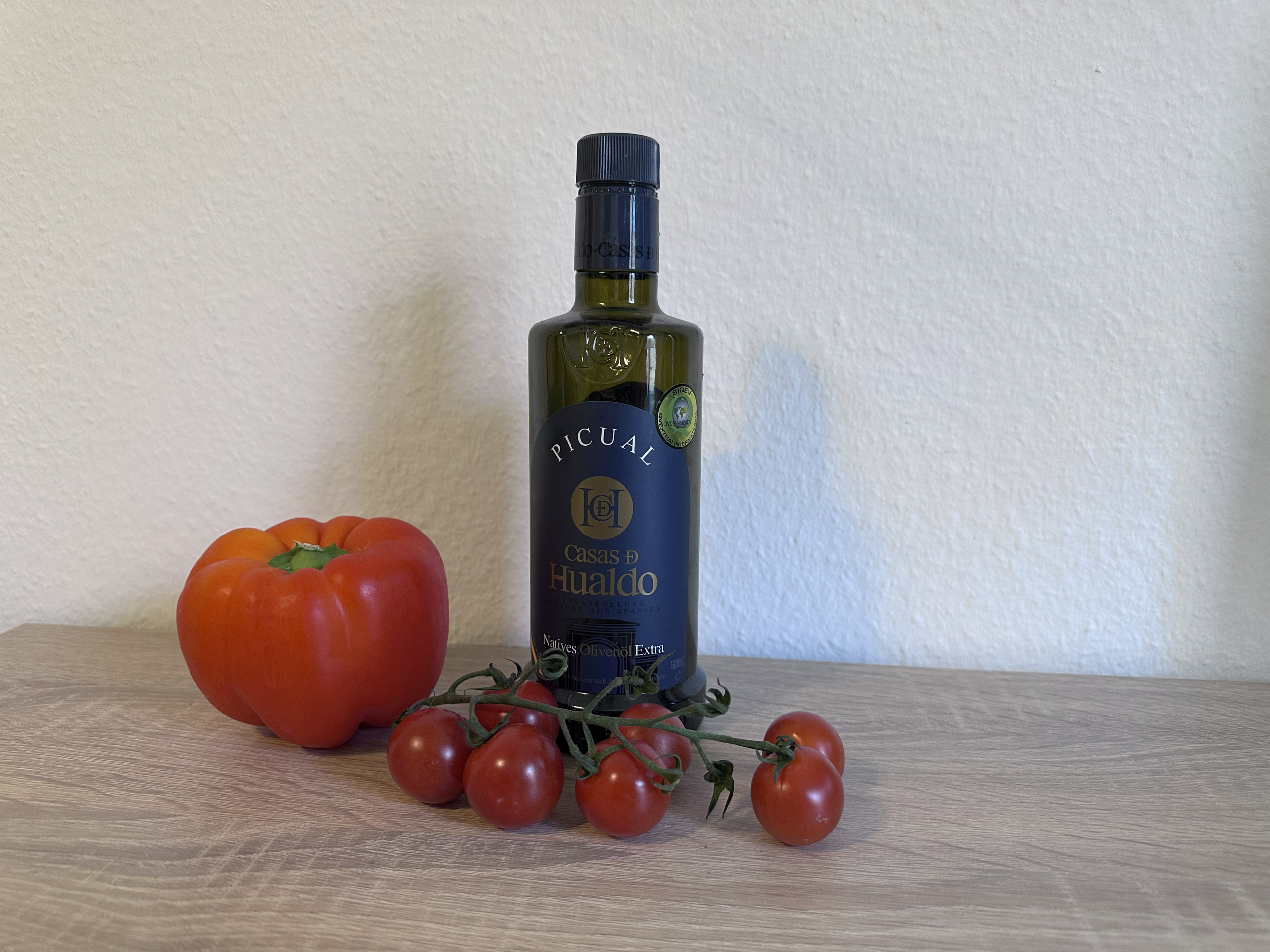 Ausschnitt eines Holztischs vor einer weißen Wand. Auf dem Tisch steht eine dunkle Flasche mit nativem Olivenöl, davor liegt eine rota Paprika sowie eine Rispe Tomaten.