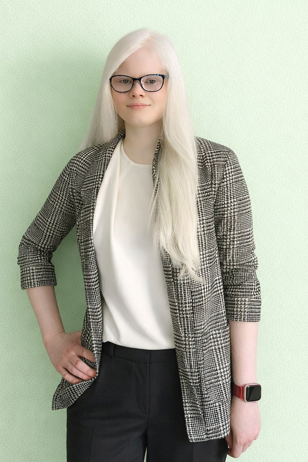 Bild von Elena Bauer, Inhaberin der Website. Man sieht einen hellgrünen Hintergrund, vor dem eine junge Frau steht. Sie hat weiße Haare und lächelt, ihre Brille ist blau, ihr Oberteil ist weiß, darüber trägt sie eine graue, karierte Jacke. Außerdem trägt sie eine Uhr mit rotem Armband und eine schwarze Anzugshose. Ihren Arm hat sie in ihre Seite gestemmt.