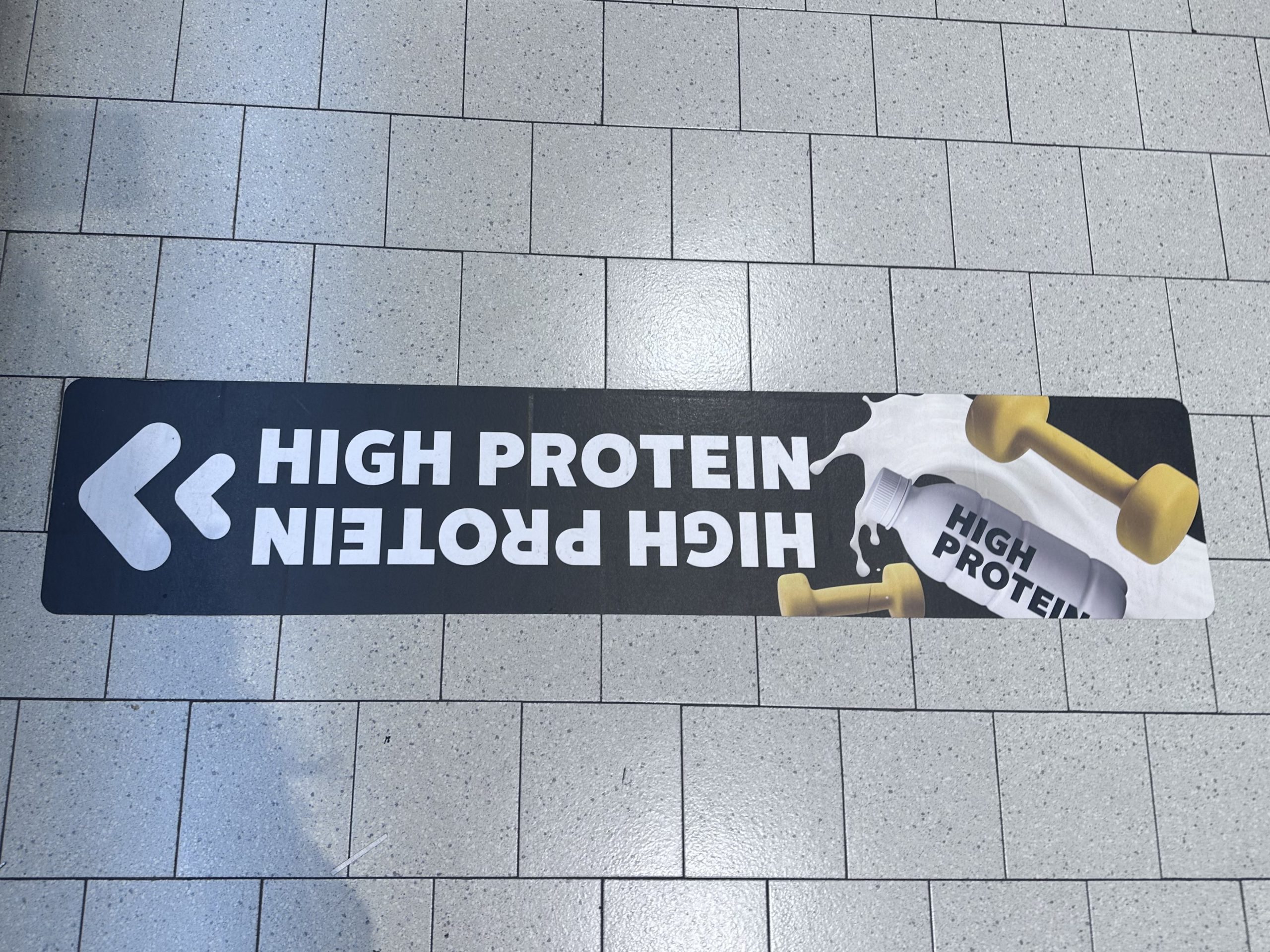 Fliesenboden in einem Supermarkt. Auf den beigen Fliesen klebt ein großer schwarzer Pfeil mit der Aufschrift "High Protein" und einer Abbildung mit zwei Hanteln und einem Proteinshake. Der Pfeil zeigt auf das Regal mit den High Protein-Produkten, welches auf dem Bild nicht sichtbar ist.