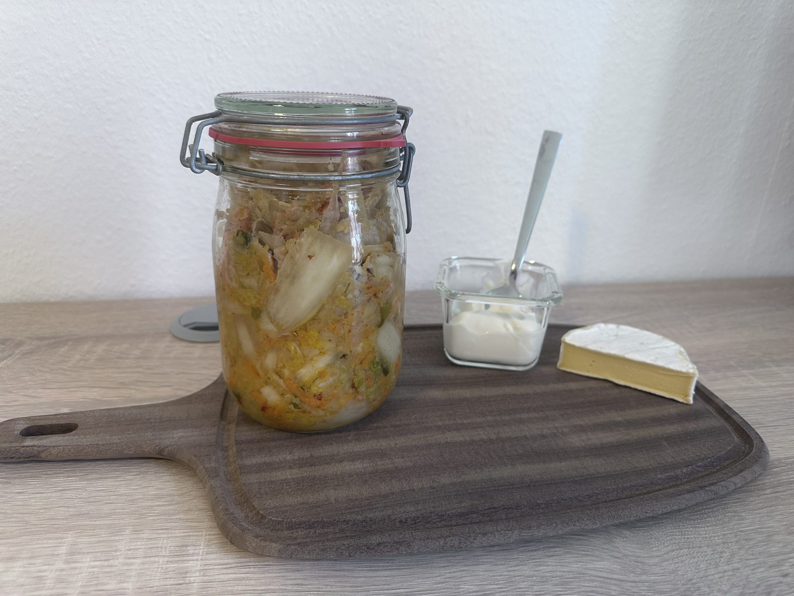 Holztisch, auf dem ein dunkles Vesperbrett aus Holz liegt. Darauf steht links ein großes Bügelglas mit Kimchi, in der Mitte steht eine kleine, eckige Glasschale mit Joghurt und einem Esslöffel, rechts liegt ein halber Camembert. Im Hintergrund ist eine weiße Wand sichtbar.