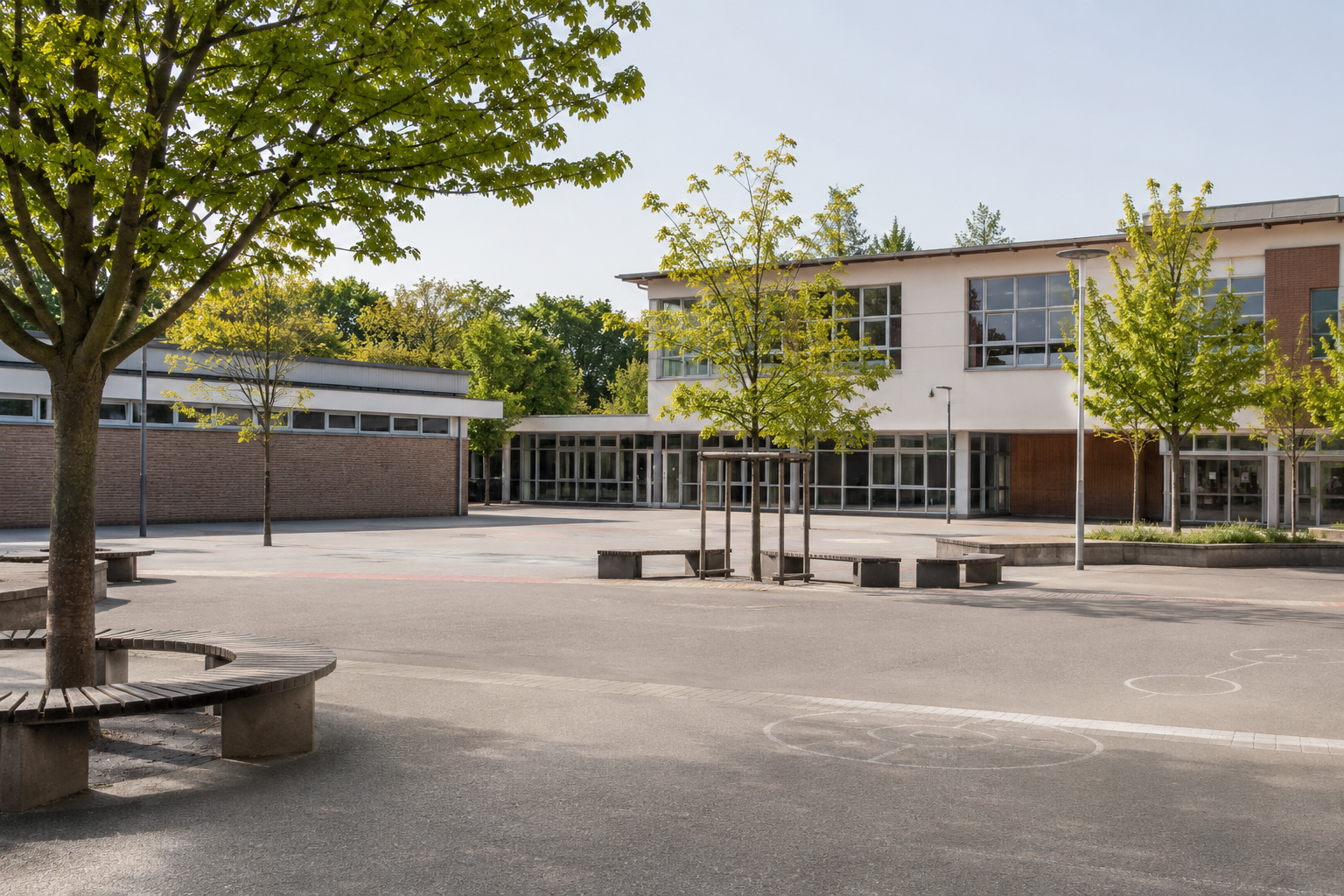 Bild eines menschenleeren Pausenhofs. Zu sehen sind im Hintergrund Gebäude, der Pausenhof selbst verfügt über einige Bäume und Sitzgelegenheiten